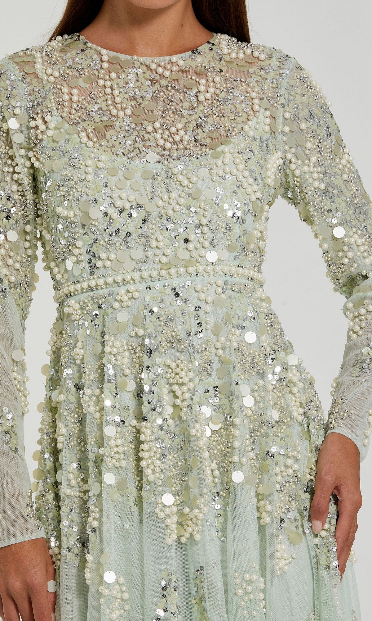 A woman models the Mac Duggal 94215 Long Formal Dress, a long-sleeved, light green evening gown embellished with sequins and beads of varying sizes, standing against a plain background.